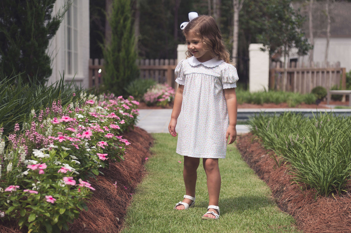 A young girl wearing a classic dress with a Peter Pan collar and puffed sleeves walks along a garden path lines with blooming pink and white flowers.  She wears white sandals and a matching white bow in her curly hair, embodying timeless Southern children