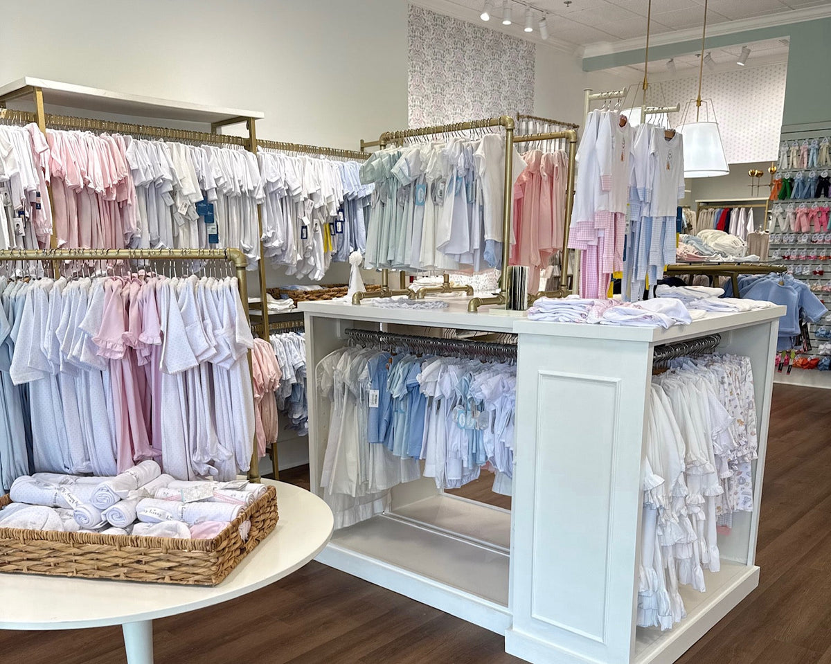 A young girl with dark braided hair and sunglasses on her head sits on a wooden ledge outdoors, holding a lemon and smiling. She is wearing a pastel floral swimsuit, surrounded by tropical greenery. A woven basket filled with citrus fruits and a glass pit